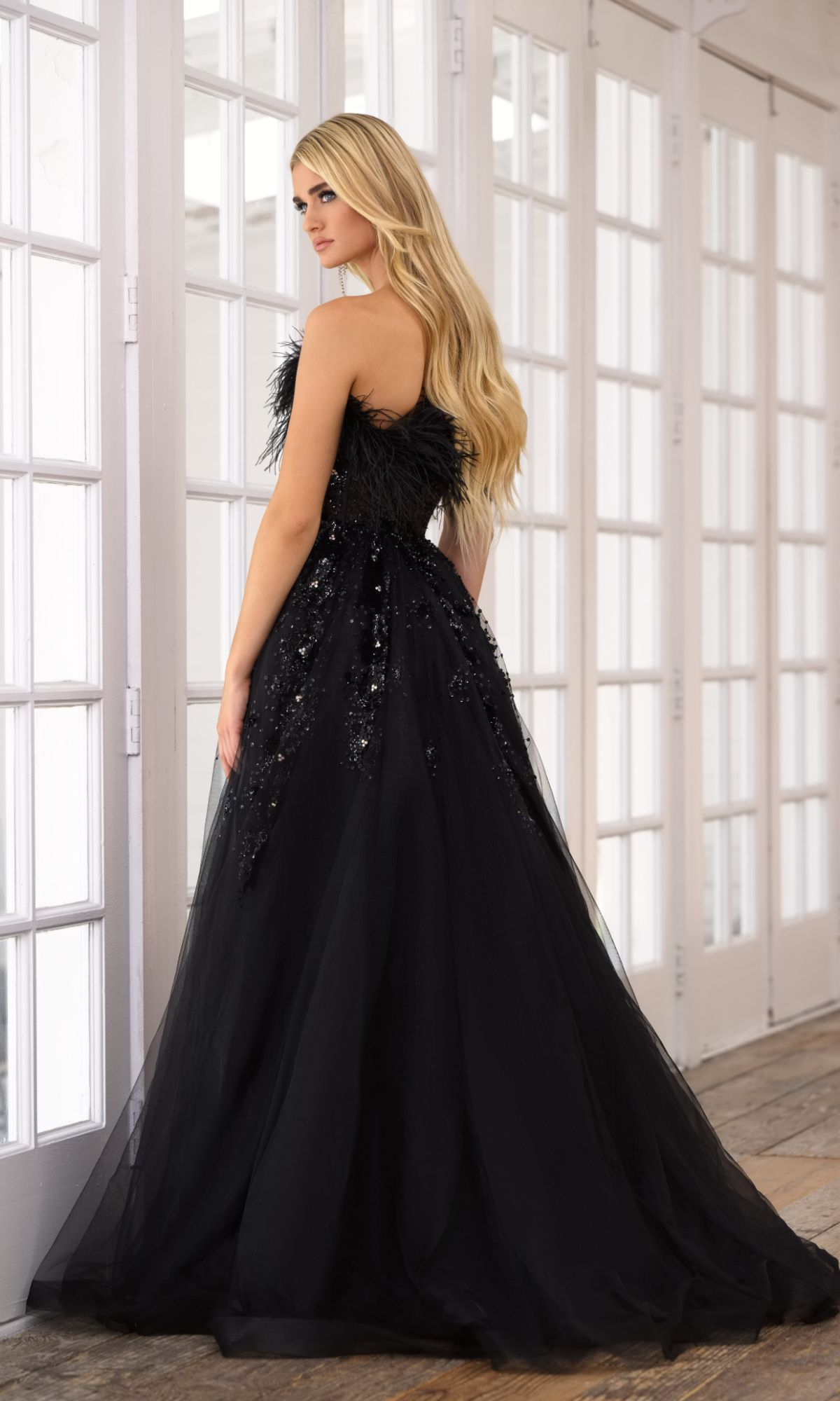 A woman in the Feather Long Prom Ball Gown: Ava Presley 39232, featuring feather trim and sequin details, stands indoors by tall white-framed windows, facing slightly away from the camera.